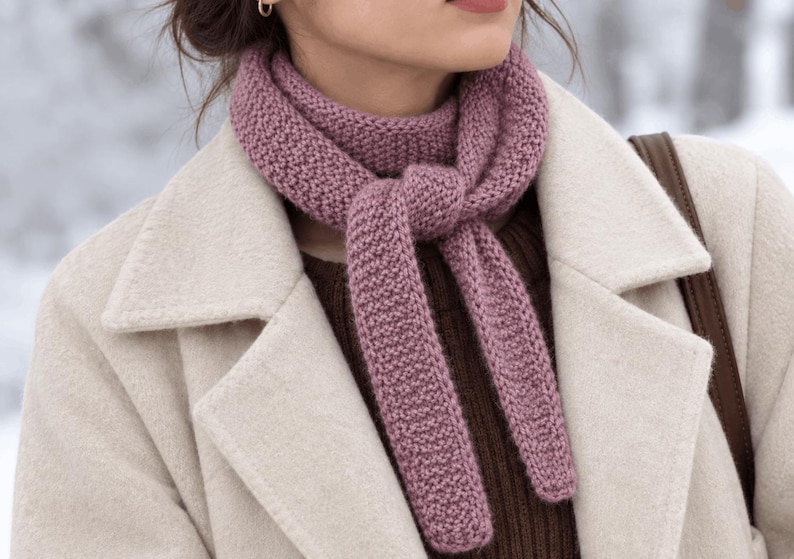 May include: A mauve knitted scarf with a textured garter stitch pattern, tied in a simple knot around the neck. The scarf is worn over a dark brown ribbed sweater and a cream-colored wool coat with wide lapels and a brown shoulder strap.