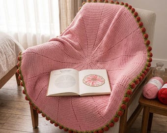 Op de afbeelding: Een ronde, roze gehaakte deken met een radiaal steekpatroon en een decoratieve rand van kleine, rode rozenknopjes met groene steeltjes. Een open instructieboek met diagrammen van het ontwerp ligt in het midden van het textielstuk.