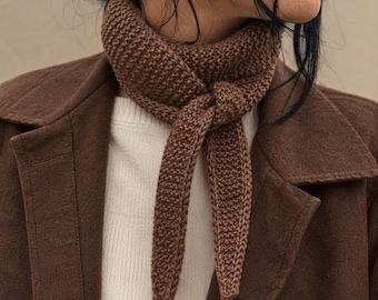 Può includere: Un fazzoletto da collo lavorato a maglia marrone con un motivo a legaccio testurizzato, annodato con un semplice nodo sul davanti. Il fazzoletto è indossato sopra un maglione a coste color crema e sotto un cappotto in misto lana marrone scuro.