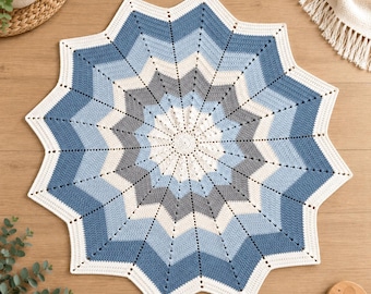Op de afbeelding: Een ronde gehaakte deken met een stervormig patroon en concentrische ringen van wit, lichtblauw, mediumblauw en grijs garen. Het ontwerp straalt vanuit een centraal punt uit naar twaalf duidelijke punten langs de buitenrand.