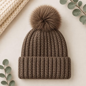 Pu&ograve; includere: Un berretto in maglia marrone scuro caratterizzato da una spessa trama a coste e un grande e soffice pon-pon in pelliccia sintetica sulla parte superiore. Il cappello presenta un ampio risvolto piegato alla base, che conferisce un aspetto strutturato.