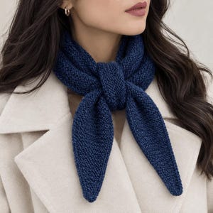 May include: A dark blue knitted scarf with a textured garter stitch pattern tied in a simple knot around the neck. The scarf features pointed ends and is worn over a light beige coat. The knit construction is visible throughout the fabric.