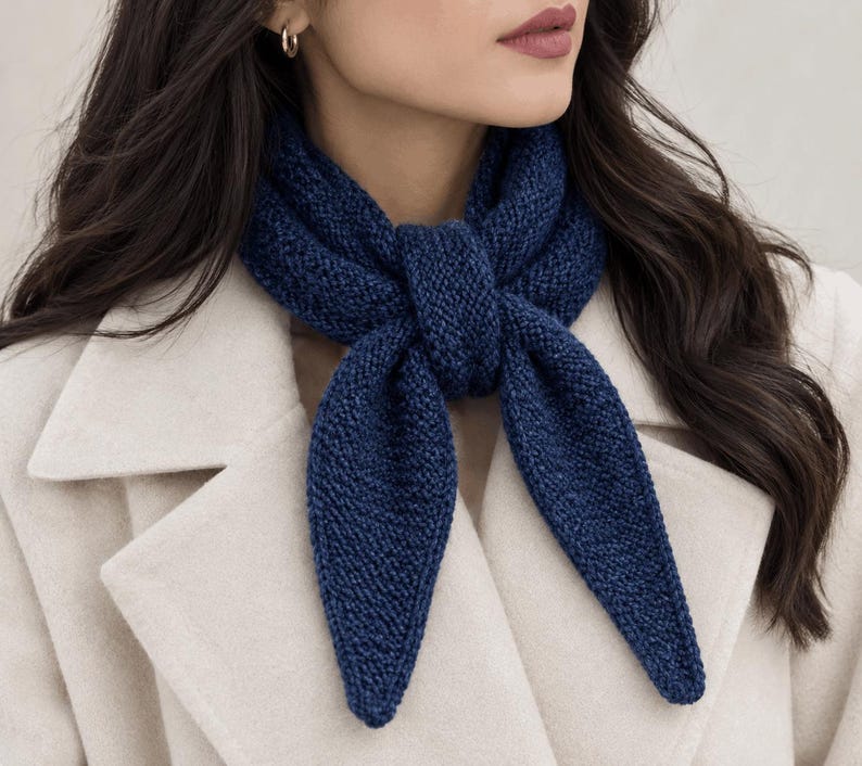 May include: A dark blue knitted scarf with a textured garter stitch pattern tied in a simple knot around the neck. The scarf features pointed ends and is worn over a light beige coat. The knit construction is visible throughout the fabric.