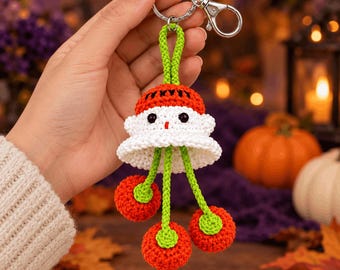 Op de afbeelding: Een gehaakte sleutelhanger met een wit, spookachtig figuurtje met zwarte ogen en een rode muts, opgehangen aan een groen koord. Drie kleine, ronde, rode gehaakte bolletjes hangen aan groene koordjes onderaan het figuurtje aan een zilveren metalen ring.