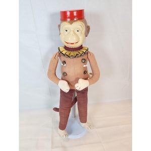 May include: A vintage monkey doll with a painted face, wearing a red hat, brown jacket, and maroon pants. The doll has a ruffled collar and button details. The monkey is standing on a white base.