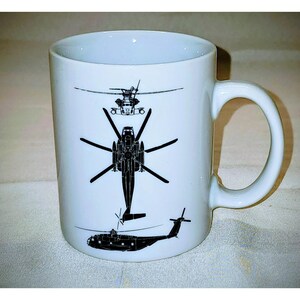 Pode incluir: Caneca de cerâmica branca com ilustrações pretas de helicópteros. O design inclui vistas superior, frontal e lateral da aeronave. A caneca tem uma alça curva e um acabamento brilhante.