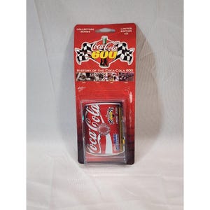 Op de afbeelding: Een Coca-Cola 600 verzamel-cd in een rode en transparante plastic verpakking. De verpakking toont het Coca-Cola logo, geblokte vlaggen en de tekst "History of the Coca-Cola 600". De cd is een limited edition.