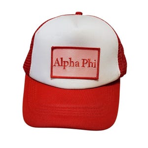 May include: A red and white trucker hat with a red peak and mesh back. The hat has a white front panel with the embroidered text "Alpha Phi" in red within a rectangular patch.
