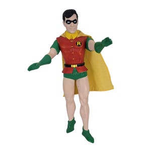Puede incluir: Una figura de acción de Robin con cabello negro, antifaz negro, túnica roja con emblema R amarillo, capa amarilla, guantes verdes, pantalones cortos verdes y botas verdes. La figura tiene cuello amarillo, cinturón negro con hebilla amarilla y piernas desnudas.
