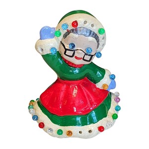May include: A ceramic Christmas figurine of a woman wearing a green coat, red skirt, and a white hat. The hat and the hem of the skirt are decorated with colorful lights. The woman is wearing glasses and has a smiling expression.
