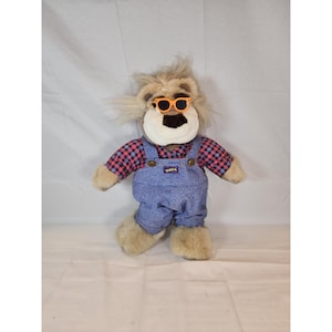 Puede incluir: Un león de peluche con gafas de sol naranjas, una camisa a cuadros rojos y negros y un peto vaquero azul. El peto tiene un pequeño parche con la palabra "QUEEN". El león tiene una melena y patas beiges.