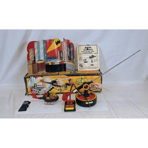 Może przedstawiać: Zestaw zabawek vintage Batcopter Batman firmy Remco, zawierający czarno-czerwony helikopter z przezroczystą kopułą, bazę sterującą z długą metalową anteną, kartonowe tło miejskie oraz instrukcję obsługi w oryginalnym pudełku.