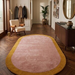 Puede incluir: Alfombra ovalada con centro rosa empolvado y borde amarillo mostaza. La alfombra est&aacute; colocada sobre un suelo de madera oscura en un pasillo. Aporta un toque de color al espacio de tonos neutros.