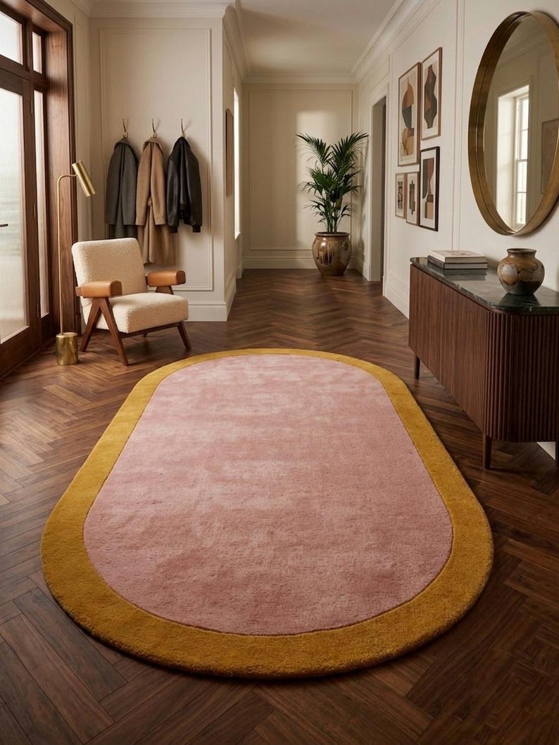 Puede incluir: Alfombra ovalada con centro rosa empolvado y borde amarillo mostaza. La alfombra est&aacute; colocada sobre un suelo de madera oscura en un pasillo. Aporta un toque de color al espacio de tonos neutros.
