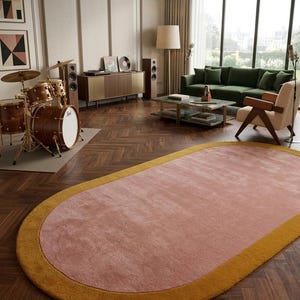 Puede incluir: Una alfombra ovalada con un centro rosa y un borde amarillo dorado. Se encuentra sobre un suelo de madera oscura en un sal&oacute;n. La habitaci&oacute;n tambi&eacute;n cuenta con un sof&aacute; verde, una bater&iacute;a y un mueble de estilo moderno de mediados de siglo.