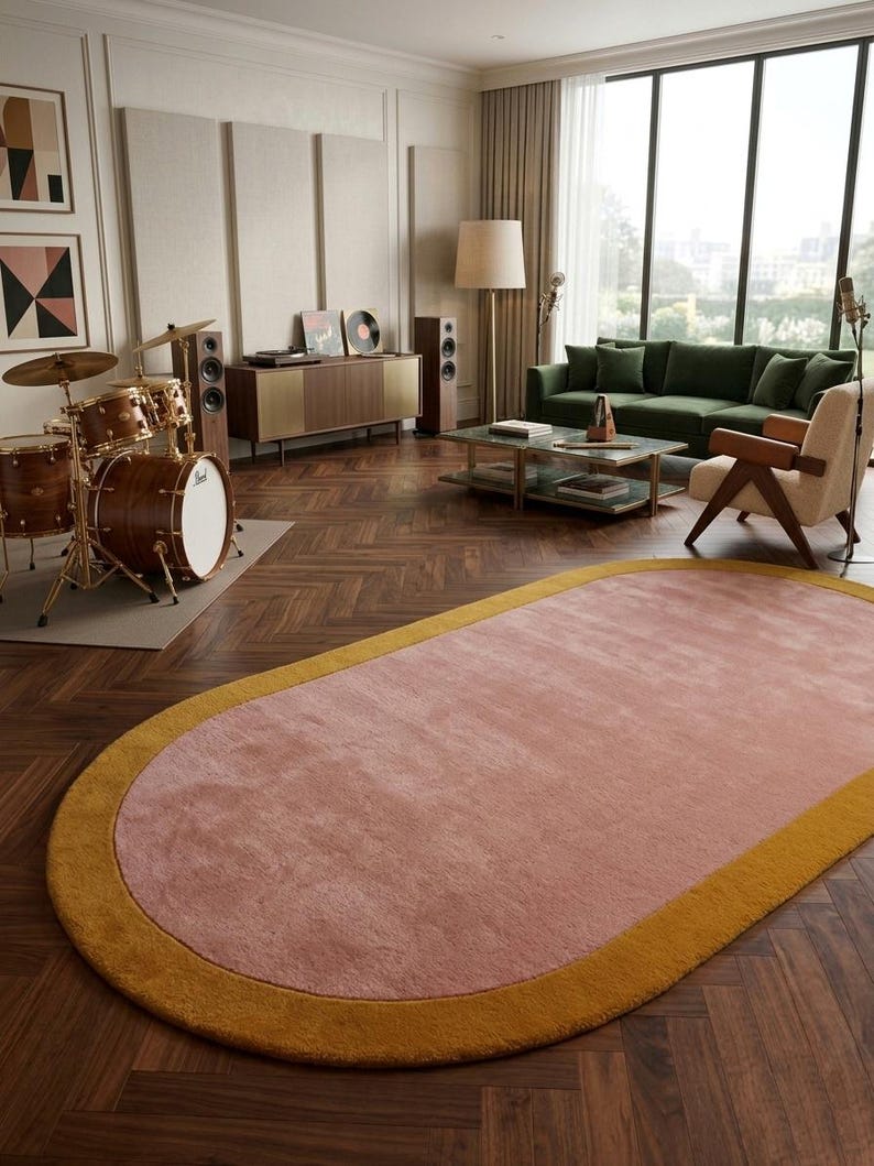 Puede incluir: Una alfombra ovalada con un centro rosa y un borde amarillo dorado. Se encuentra sobre un suelo de madera oscura en un sal&oacute;n. La habitaci&oacute;n tambi&eacute;n cuenta con un sof&aacute; verde, una bater&iacute;a y un mueble de estilo moderno de mediados de siglo.
