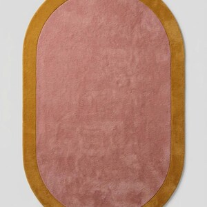 Puede incluir: Una alfombra ovalada con un centro rosa empolvado y un borde amarillo dorado. La alfombra tiene una textura suave y afelpada, y la combinaci&oacute;n de colores crea una est&eacute;tica c&aacute;lida. Adecuada para salones y dormitorios, mide unos 150 cm x 90 cm.