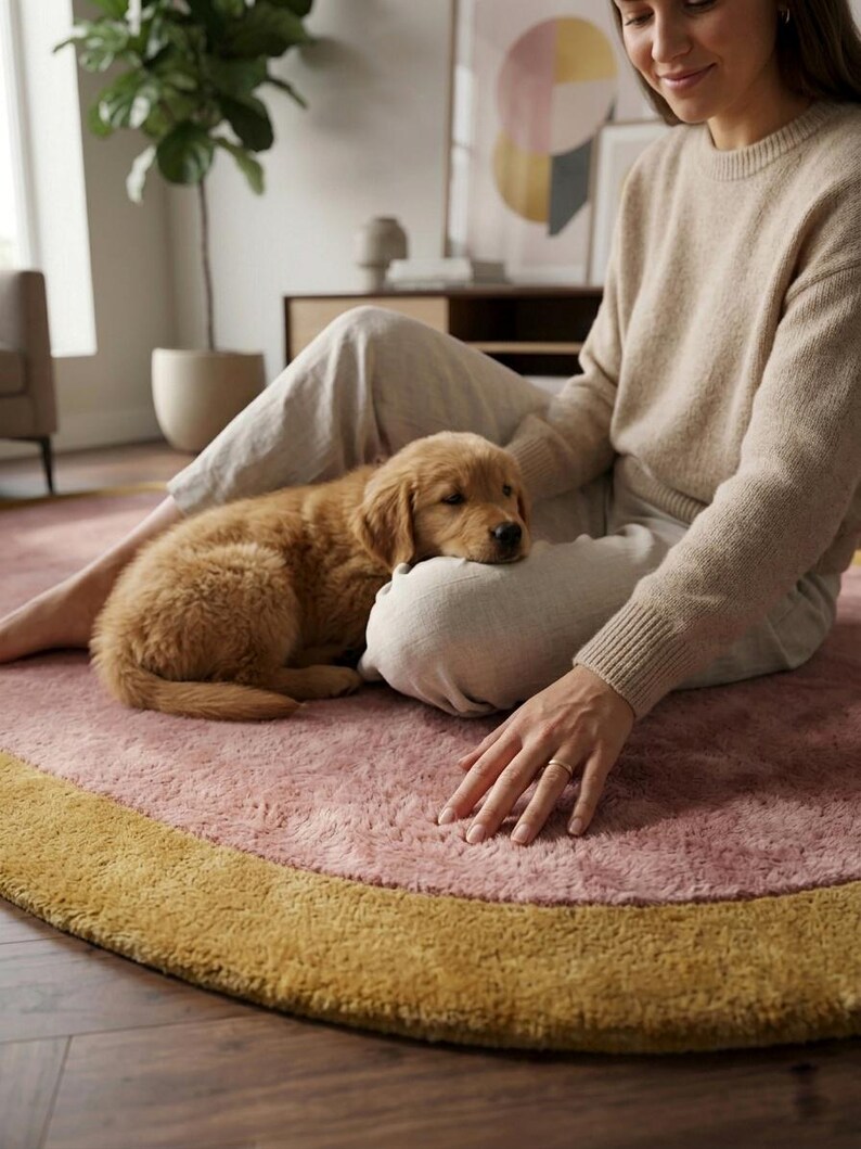 Puede incluir: Un cachorro golden retriever descansa sobre una alfombra rosa con borde amarillo. La alfombra est&aacute; en un sal&oacute;n, con una persona que lleva un su&eacute;ter y pantalones beiges. Una planta de interior es visible en el fondo.