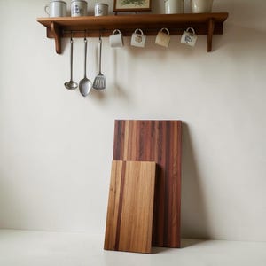 Könnte beinhalten: Zwei Holzschneidebretter, eines rechteckig und eines kleiner, werden vor einer weißen Wand präsentiert. Ein Holzregal darüber enthält Tassen und Küchenutensilien. Die Bretter haben eine natürliche Holzmaserung.
