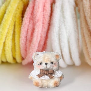 May include: A miniature teddy bear, handmade with a white lace collar and a brown bow. The bear has rosy cheeks and a white lace headpiece. Soft, colourful yarn in yellow, pink, white, and beige are in the background.