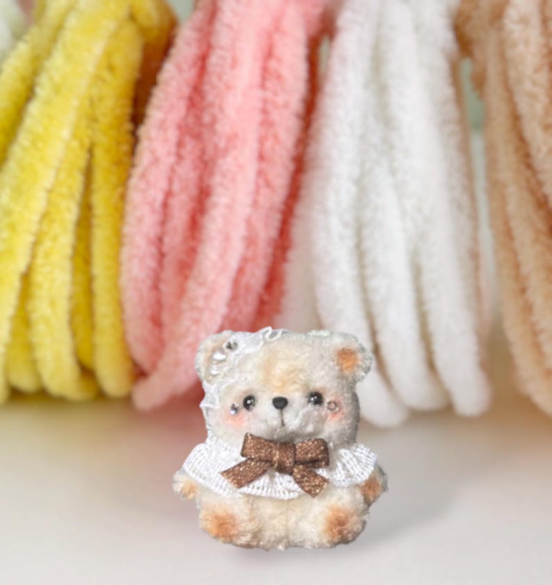 May include: A miniature teddy bear, handmade with a white lace collar and a brown bow. The bear has rosy cheeks and a white lace headpiece. Soft, colourful yarn in yellow, pink, white, and beige are in the background.