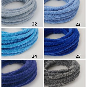 May include: Six plush hair scrunchies in shades of blue and grey are displayed. Each circular scrunchie is numbered from 22 to 27, indicating different colour options. The fabric has a soft, textured appearance.