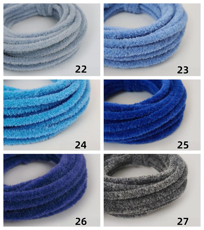 May include: Six plush hair scrunchies in shades of blue and grey are displayed. Each circular scrunchie is numbered from 22 to 27, indicating different colour options. The fabric has a soft, textured appearance.
