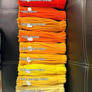 May include: Ten bundles of fuzzy pipe cleaners arranged in a vertical gradient from dark orange to pale yellow. Each bundle is labeled with a white tag and text: YL01 Persimmon Orange, YL02 Carrot, YL03 Ginger Bread, YL04 Orange, YL05 Golden Yellow, YL06 Mango Yellow, YL07 Lemon Yellow, YL08 Gardenia Yellow, YL09 Butter Yellow, YL10 Primorse Yellow.