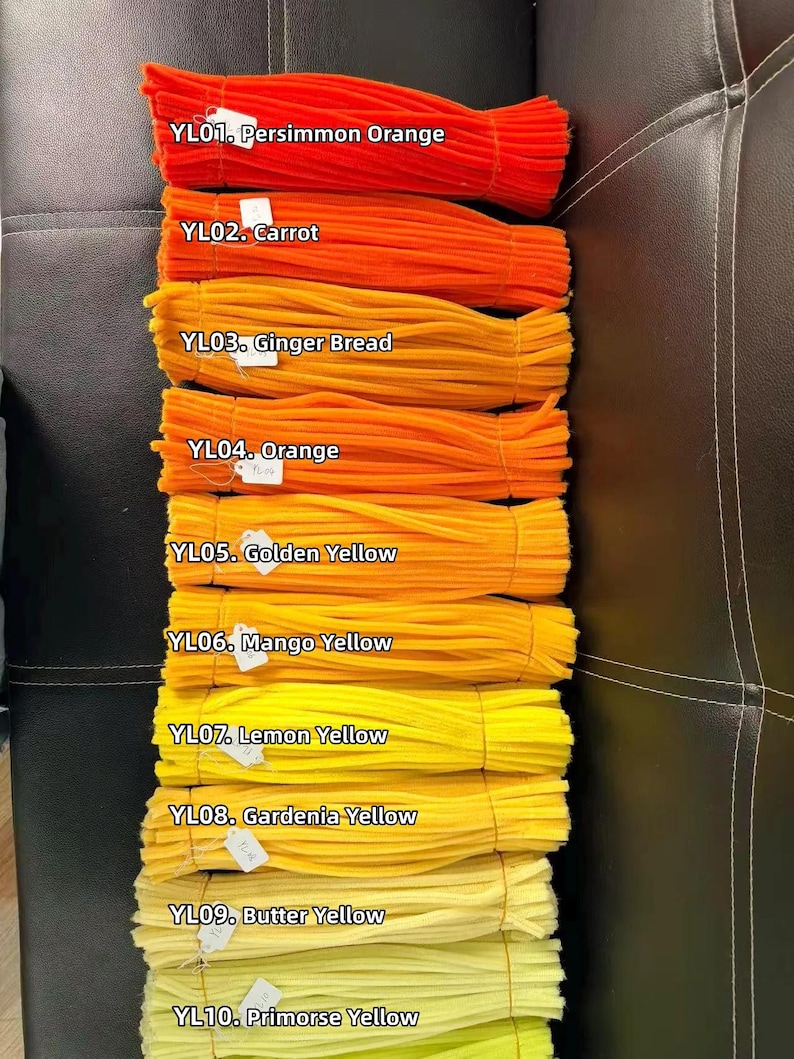May include: Ten bundles of fuzzy pipe cleaners arranged in a vertical gradient from dark orange to pale yellow. Each bundle is labeled with a white tag and text: YL01 Persimmon Orange, YL02 Carrot, YL03 Ginger Bread, YL04 Orange, YL05 Golden Yellow, YL06 Mango Yellow, YL07 Lemon Yellow, YL08 Gardenia Yellow, YL09 Butter Yellow, YL10 Primorse Yellow.