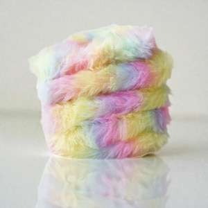 May include: A stack of soft, fluffy fabric in pastel shades. The material is a blend of yellow, pink, purple, and light blue, producing a rainbow effect. The fabric looks like faux fur or a similar plush texture, with a subtle sheen.
