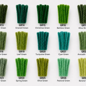 May include: A collection of fifteen bundles of fuzzy pipe cleaners arranged in a grid, each labeled with a code and color name ranging from Moss Green GR13 to Banana Green GR27, showcasing various shades of green in a textured, fibrous material.