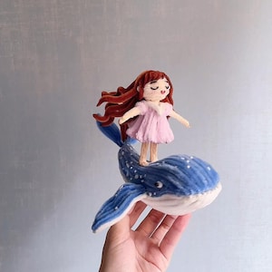 Peut inclure: Une figurine en feutrine artisanale repr&eacute;sentant un petit personnage aux longs cheveux bruns v&ecirc;tu d'une robe rose p&acirc;le, debout sur une baleine bleue &agrave; pois blancs. La baleine poss&egrave;de des nageoires marqu&eacute;es et un ventre blanc textur&eacute;.