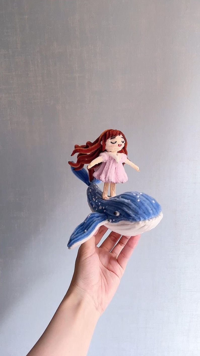Peut inclure: Une figurine en feutrine artisanale repr&eacute;sentant un petit personnage aux longs cheveux bruns v&ecirc;tu d'une robe rose p&acirc;le, debout sur une baleine bleue &agrave; pois blancs. La baleine poss&egrave;de des nageoires marqu&eacute;es et un ventre blanc textur&eacute;.
