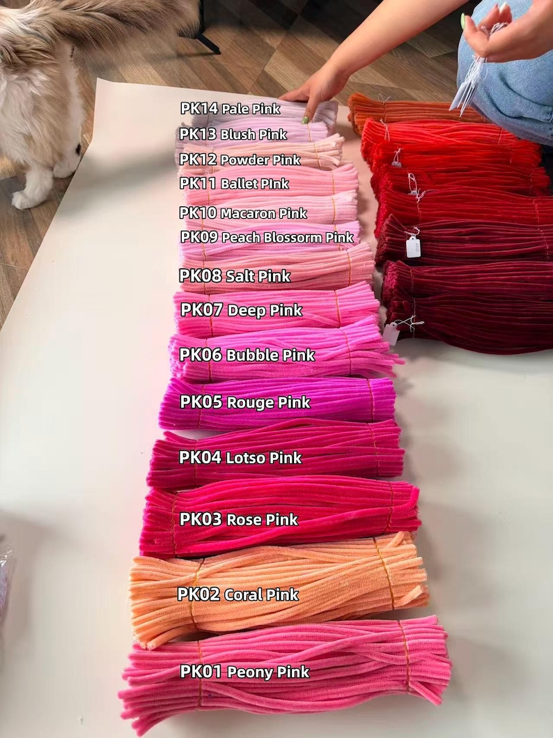May include: Bundles of fuzzy pipe cleaners arranged in a vertical column, labeled with color names from PK14 Pale Pink to PK01 Peony Pink. To the right, additional bundles of red and orange pipe cleaners are stacked in horizontal rows.
