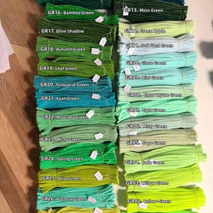May include: A collection of fuzzy pipe cleaners arranged in two columns, labeled GR01 through GR27 with specific color names ranging from Neon Green to Christmas Green, showcasing a variety of green shades including mint, olive, and emerald.