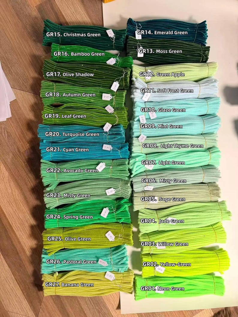 May include: A collection of fuzzy pipe cleaners arranged in two columns, labeled GR01 through GR27 with specific color names ranging from Neon Green to Christmas Green, showcasing a variety of green shades including mint, olive, and emerald.