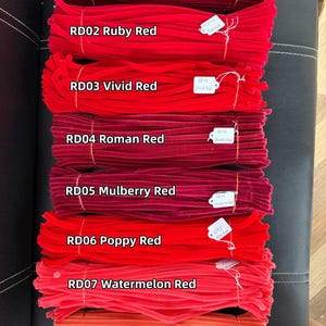 May include: Eight bundles of fuzzy pipe cleaners arranged vertically, each labeled with a white tag and text: RD01 Magenta Red, RD02 Ruby Red, RD03 Vivid Red, RD04 Roman Red, RD05 Mulberry Red, RD06 Poppy Red, RD07 Watermelon Red, and RD08 Grapefruit Red.
