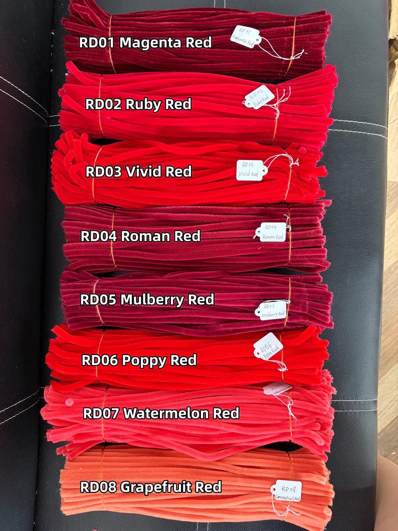 May include: Eight bundles of fuzzy pipe cleaners arranged vertically, each labeled with a white tag and text: RD01 Magenta Red, RD02 Ruby Red, RD03 Vivid Red, RD04 Roman Red, RD05 Mulberry Red, RD06 Poppy Red, RD07 Watermelon Red, and RD08 Grapefruit Red.