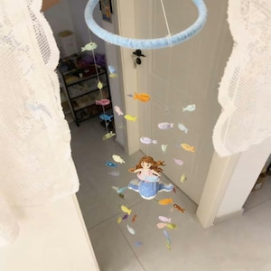 Peut inclure: Un mobile suspendu compos&eacute; d'un cadre circulaire bleu clair avec plusieurs fils de petits poissons color&eacute;s. Une figurine centrale de sir&egrave;ne aux cheveux bruns et &agrave; la queue bleue est assise sur une base en forme de nuage blanc.