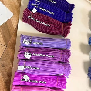 May include: Bundles of fuzzy pipe cleaners in various shades of purple and pink, labeled with white tags: PP01 Deep purple, PP02 Indigo Purple, PP03 Berry Purple, PP05 Wisteria, PP06 Dark Plum Purple, PP07 Violet, PP08 Fuchsia Violet, and PP09 Pink Lavender.