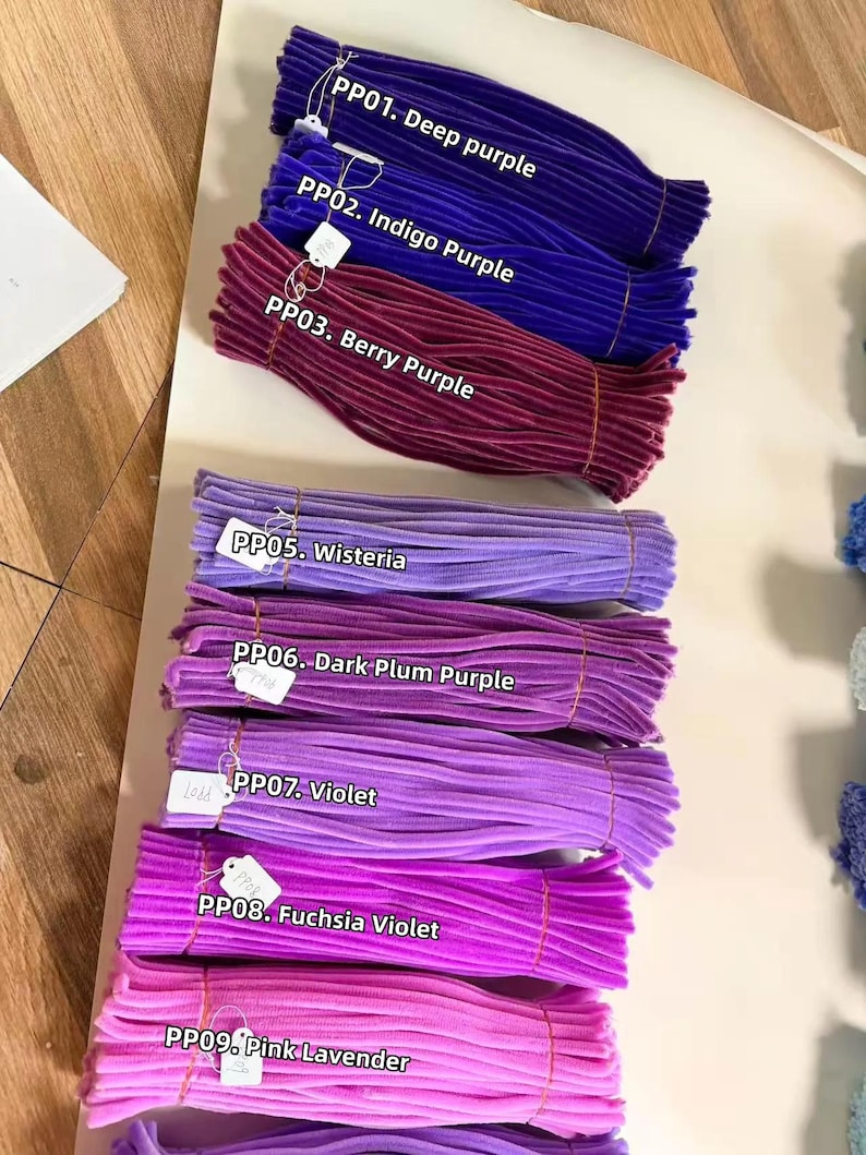 May include: Bundles of fuzzy pipe cleaners in various shades of purple and pink, labeled with white tags: PP01 Deep purple, PP02 Indigo Purple, PP03 Berry Purple, PP05 Wisteria, PP06 Dark Plum Purple, PP07 Violet, PP08 Fuchsia Violet, and PP09 Pink Lavender.