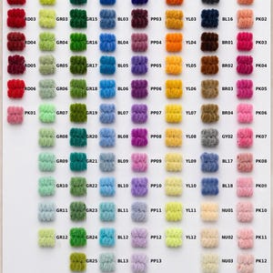 May include: A color chart displaying 104 small, fuzzy, cylindrical yarn pom-poms arranged in columns and rows. Each pom-pom is labeled with a unique alphanumeric code such as RD01, GR01, or BL01. The text 104 Colours is printed in orange at the bottom left.