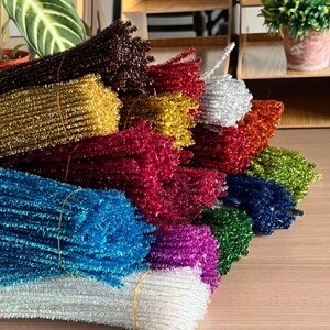 May include: A selection of colourful pipe cleaner bundles, including gold, red, blue, and green. Each bundle is secured with an elastic band. The pipe cleaners have a fuzzy, textured appearance, suitable for craft projects.