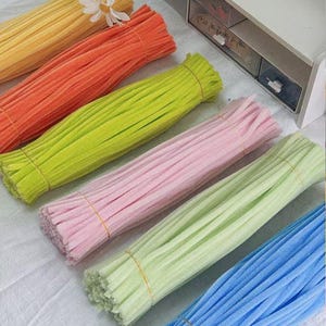 May include: Six bundles of fuzzy pipe cleaners arranged in a diagonal row. The colors include yellow, orange, lime green, light pink, pale mint green, and sky blue. Each bundle is secured with a thin rubber band around the center of the stems.