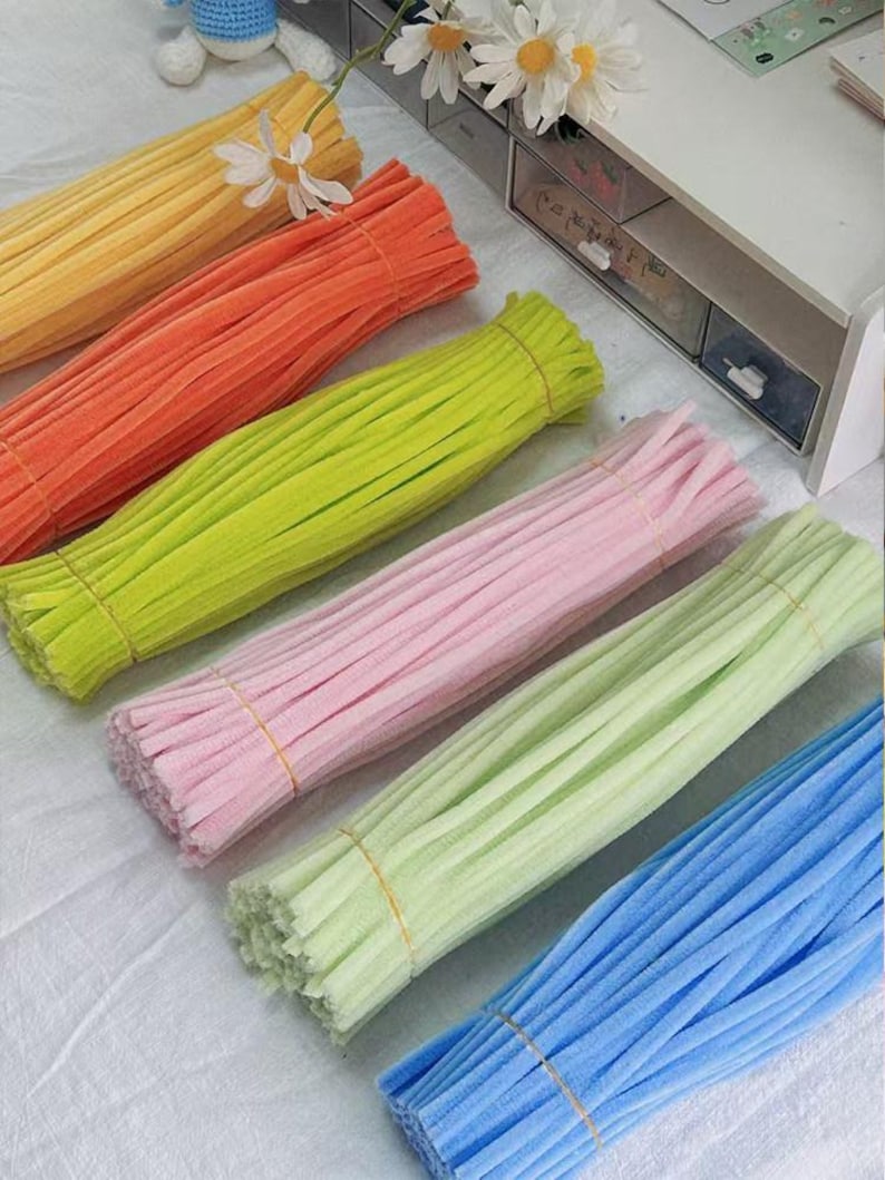 May include: Six bundles of fuzzy pipe cleaners arranged in a diagonal row. The colors include yellow, orange, lime green, light pink, pale mint green, and sky blue. Each bundle is secured with a thin rubber band around the center of the stems.