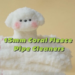 Op de afbeelding: Close-up van witte, pluizige 15 mm koraalvlies pijpenragers. De afbeelding toont een witte, zachte, hondachtige figuur met roze wangen en zwarte ogen. De tekst "15mm Coral Fleece Pipe Cleaners" is groen.