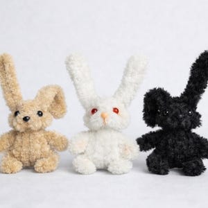 May include: Three handmade plush bunny rabbits in tan, white, and black. Each bunny has long, floppy ears and small, round bodies. The white bunny has red eyes. These soft toys are perfect for Easter or as a unique gift.