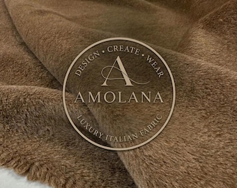Puede incluir: Una pieza de tela de piel sintética marrón con un pelo suave y texturizado. Un logotipo circular está superpuesto en el centro, con el texto DESIGN CREATE WEAR, la letra A, la palabra AMOLANA y la frase LUXURY ITALIAN FABRIC.