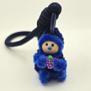 May include: A dark blue fuzzy hair tie featuring a small, plush, blue-hooded character with a light-coloured face, black eyes, and a blue nose. The character is holding a small, purple, grape-shaped charm with a green leaf accent in its hands.