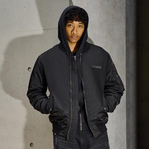 Puede incluir: Chaqueta bomber negra con cierre de cremallera, bolsillos laterales y un peque&ntilde;o parche rectangular en el pecho. La chaqueta se lleva sobre una sudadera con capucha negra y vaqueros negros. El fondo es una pared gris texturizada.
